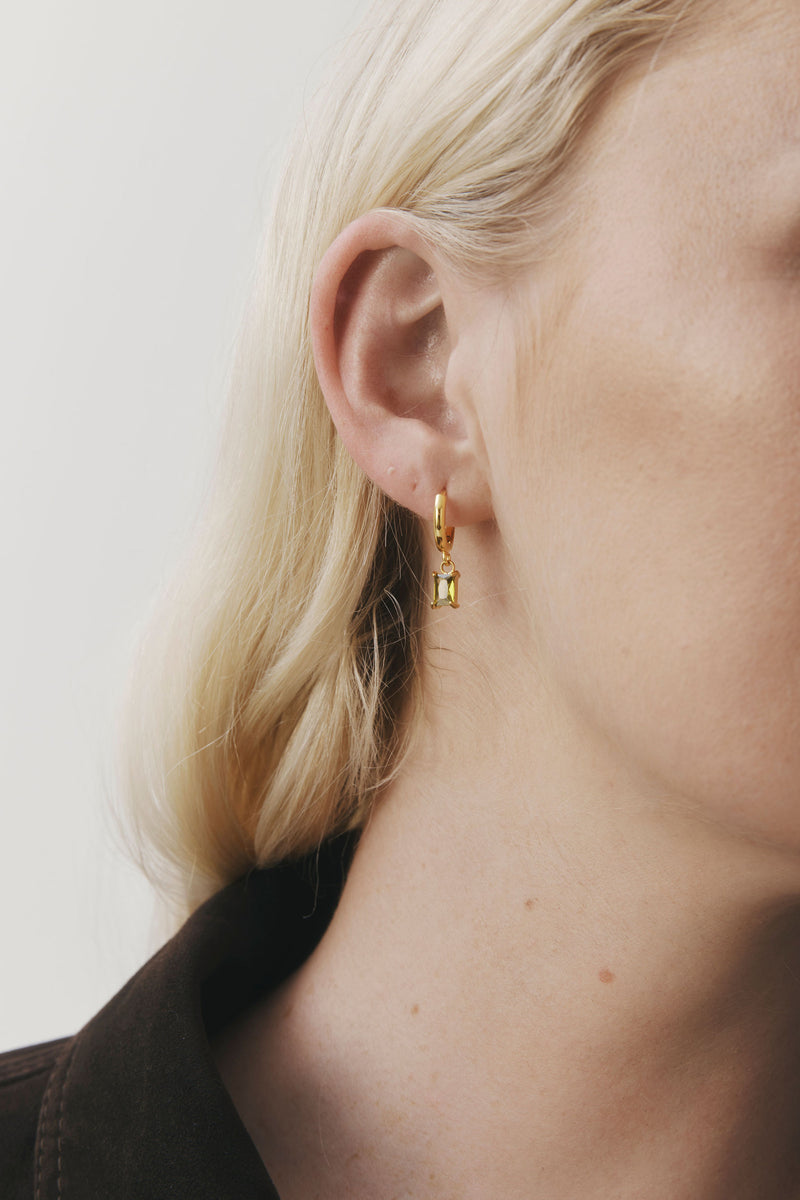 Arina Earrings by Castel & Co are gold-plated stainless steel hoop earrings featuring an olive green stone drop. Designed with a slim, elegant silhouette, they offer a lightweight feel (3g) and a Congo closure, measuring 2.2 x 0.4 cm. Crafted to catch the light with movement, they’re suitable for both everyday wear and evening styling. Hypoallergenic and waterproof.
