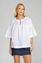 The distinguished BLAK Thea Top presents an elegant interplay of white fabric and refined black stitching. Crafted from premium cotton voile, this sophisticated piece features a gracefully gathered round neckline complemented by a tasteful front keyhole with delicate ties. The artfully designed shirt sleeves showcase meticulous shirring details, echoed in the thoughtful shirring embellishments adorning both the upper front and back. The silhouette extends precisely to mid-hip, concluding with a pristine str