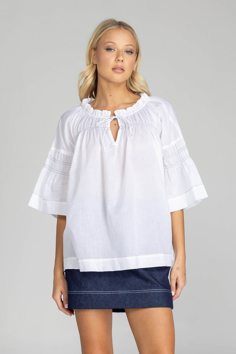 The distinguished BLAK Thea Top presents an elegant interplay of white fabric and refined black stitching. Crafted from premium cotton voile, this sophisticated piece features a gracefully gathered round neckline complemented by a tasteful front keyhole with delicate ties. The artfully designed shirt sleeves showcase meticulous shirring details, echoed in the thoughtful shirring embellishments adorning both the upper front and back. The silhouette extends precisely to mid-hip, concluding with a pristine str