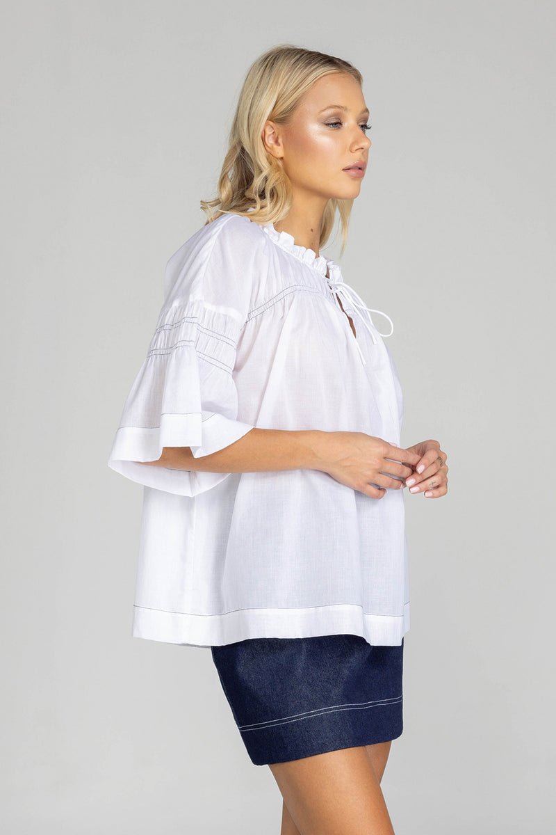 The distinguished BLAK Thea Top presents an elegant interplay of white fabric and refined black stitching. Crafted from premium cotton voile, this sophisticated piece features a gracefully gathered round neckline complemented by a tasteful front keyhole with delicate ties. The artfully designed shirt sleeves showcase meticulous shirring details, echoed in the thoughtful shirring embellishments adorning both the upper front and back. 
